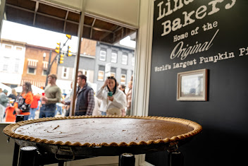 Lindsey's Bakery window display featuring a large pumpkin pie with visitors visible through the glass on the street