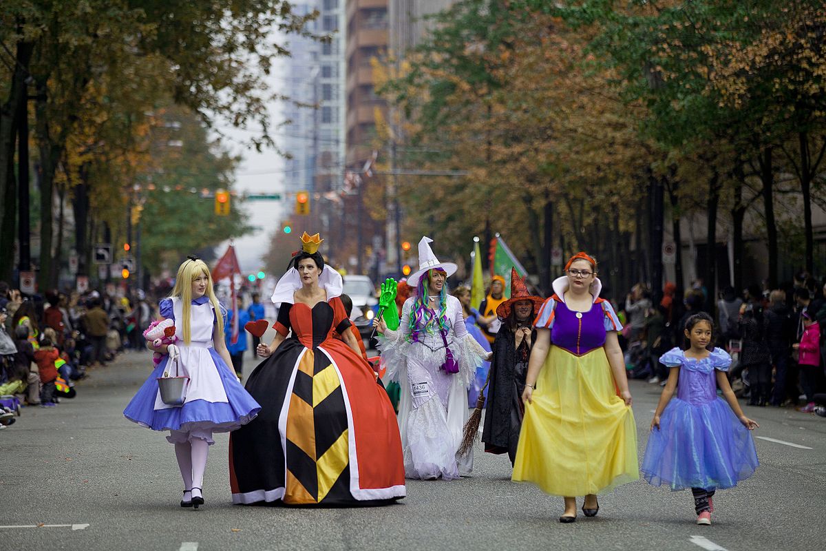 Costumed parade marchers dressed as storybook characters, including Alice and the Queen of Hearts, walk down a city street