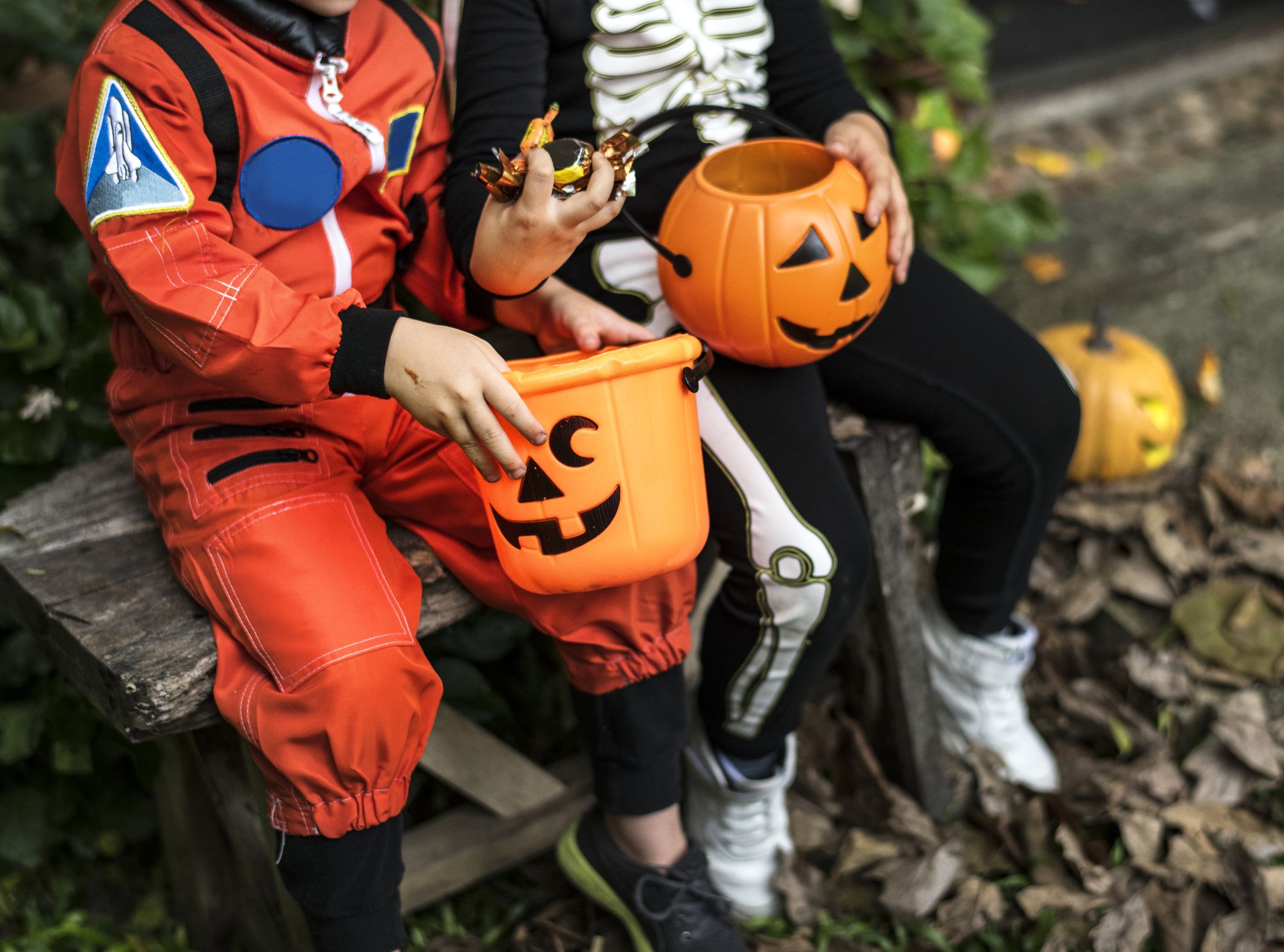 Two children in astronaut and skeleton costumes sitting on a bench holding jack-o'-lantern candy buckets